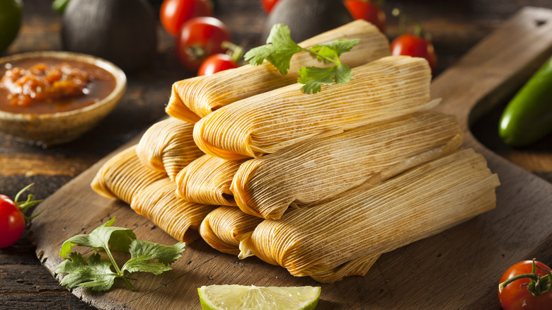 A neat pile of tamales on a pizza board surrounded by tomatoes and avocados