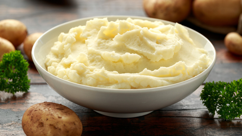 Bowl of mashed potatoes surrounded by potatoes and herbs