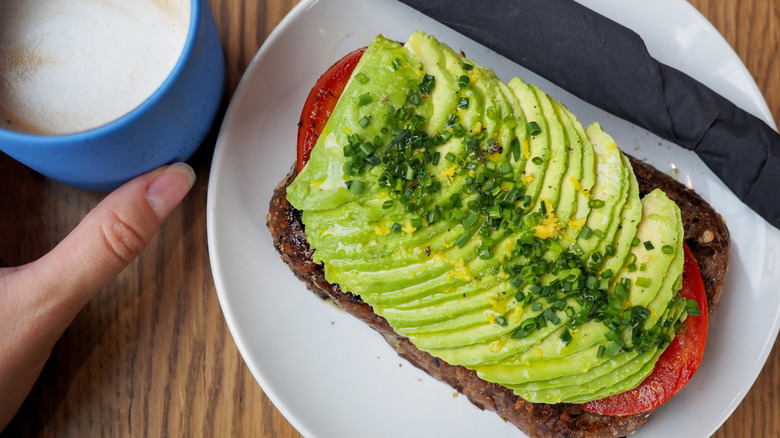 Piece of avocado toast with chives and tomatoes next to a latte.