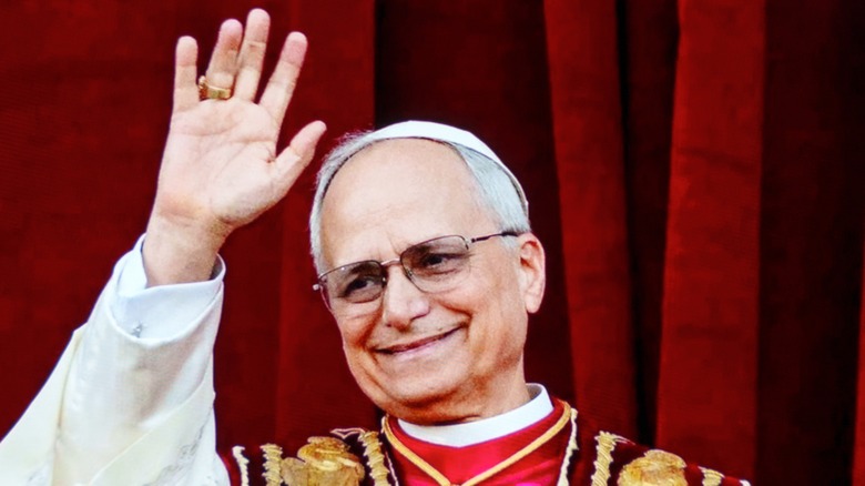 Pope Leo XIV smiles and waves with a red velvet curtain behind him