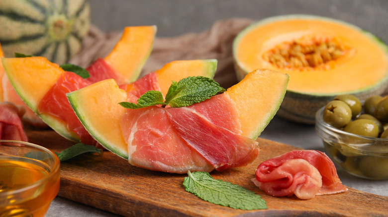 Melon and prosciutto on a wooden serving platter with mint and bowls of olives and honey.