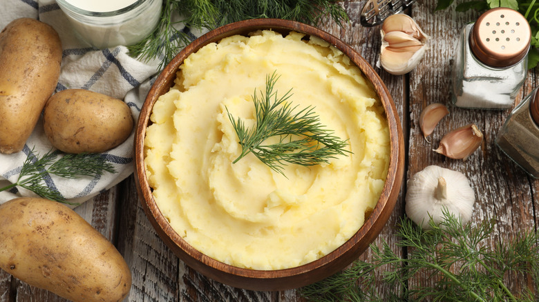 Mashed potatoes topped with a dill sprig in a wooden bowl, with whole potatoes and garlic around it