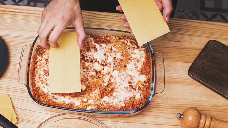 a person's hands layering lasagna with pasta sheets in a glass baking dish