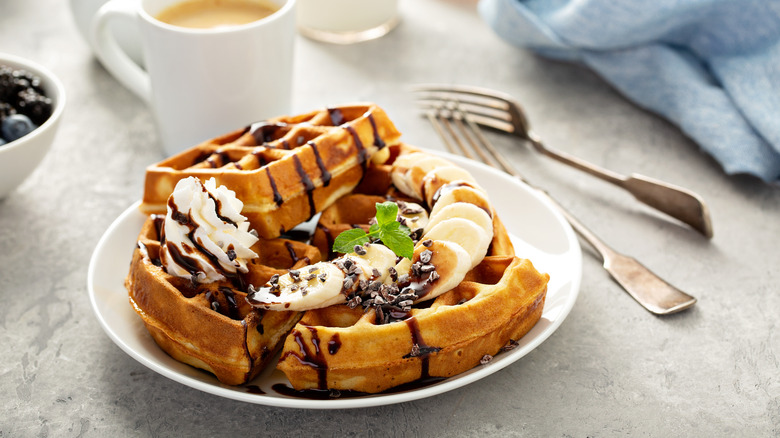 waffles with bananas, whipped cream, and chocolate sauce on a plate with a bowl of blueberries to the left, forks on the right, and a cup of coffee