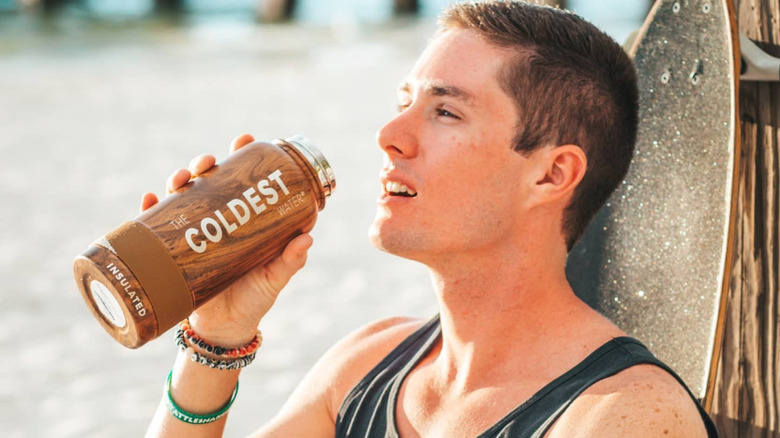 A skateboarder sits against their board while lifting a brown Coldest bottle of water to drink from