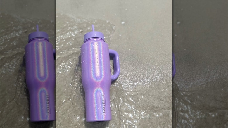 An iridescent purple Coldest water bottle lies on a sandy beach as the ocean washes over it