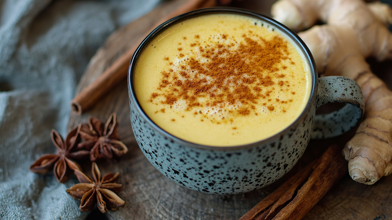 A mug of golden milk with spices sprinkled on top and whole star anise pods, whole cinnamon, and turmeric root beside the cup