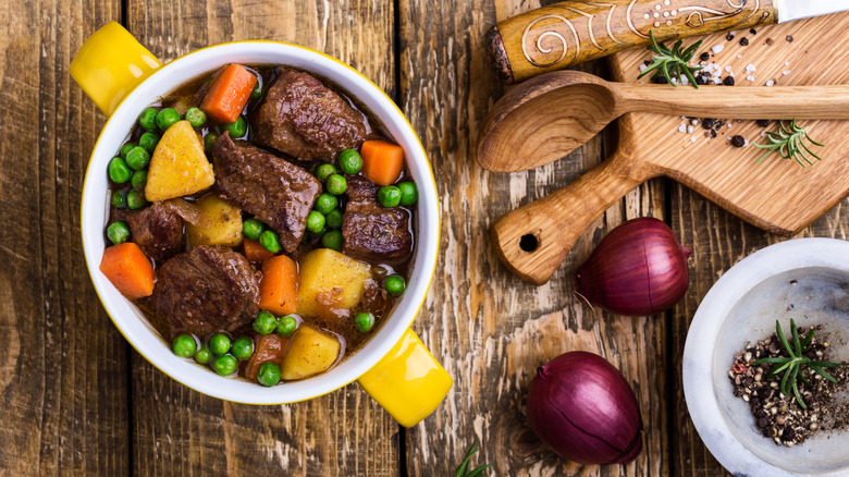 A bowl of thick beef stew with potatoes, carrots, and peas on a countertop surrounded by red onions, a bowl of crushed black pepper, cooking utensils, and a cutting board with a wooden spoon, knife, and spices on top.