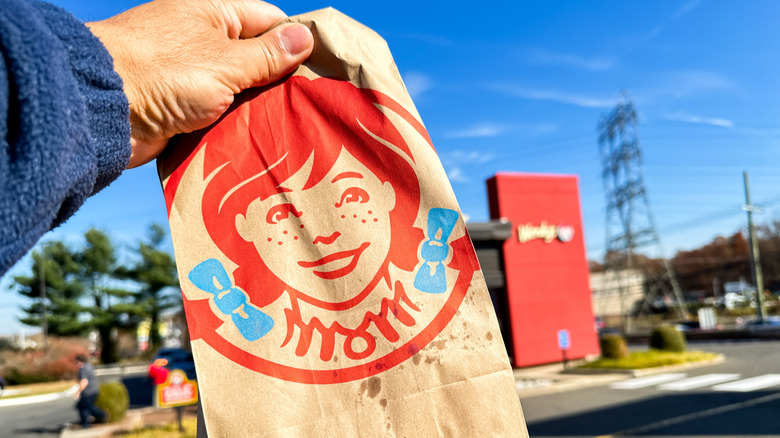 A close up of the Wendy's logo on a bag of food held in front of a Wendy's.