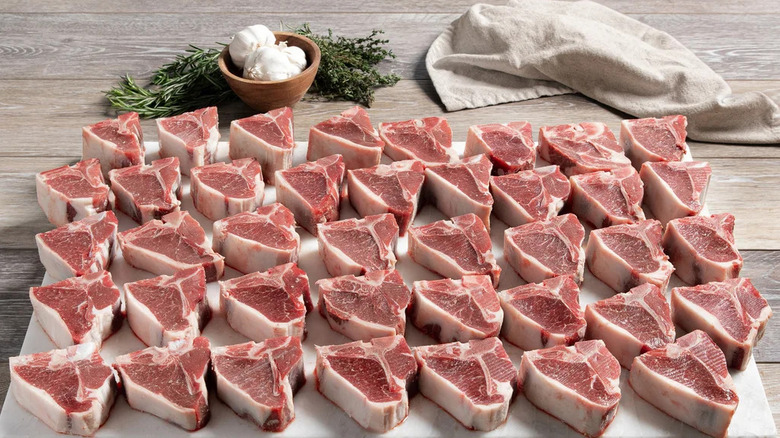 a whole mess of raw lamb chops on a large cutting board next to fresh herbs and garlic