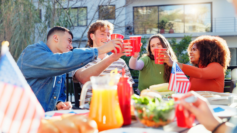 A group of friends sit around an outdoor barbecue with American flags, cheersing from red Solo cups