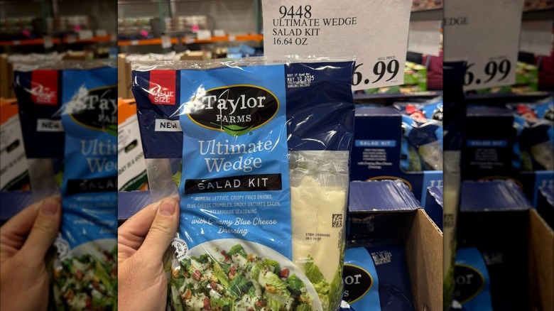 A hand holds up a bag of Taylor Farms Ultimate Wedge Salad Kit at Costco