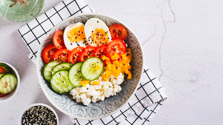 A bowl of cottage cheese has salad toppings, including tomatoes, cucumbers, diced peppers, boiled eggs, and sesame seeds