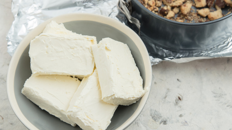 Four rectangular blocks of white cream cheese in a white bowl next to a round baking pan with cookie crumbles in it