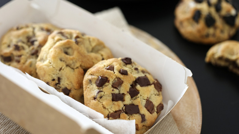 Three puffy chocolate chunk cookies in a rectangular white paper container
