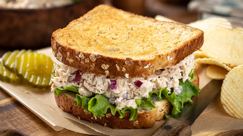 tuna salad sandwich on cutting board next to pickle slices and potato chips