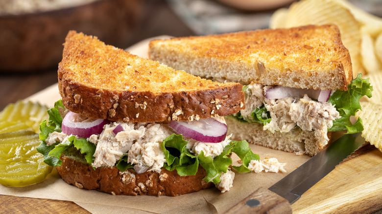 Tuna salad sandwich on a cutting board next to pickle slices and potato chips