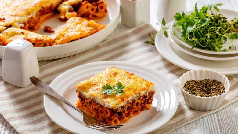 A plate of pastitsio next to fresh herbs, a bowl of black pepper, and a casserole dish.