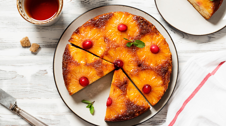 A pretty pineapple upside down cake with cherries has a sliced removed and is topped with fresh mint beside a cup of tea