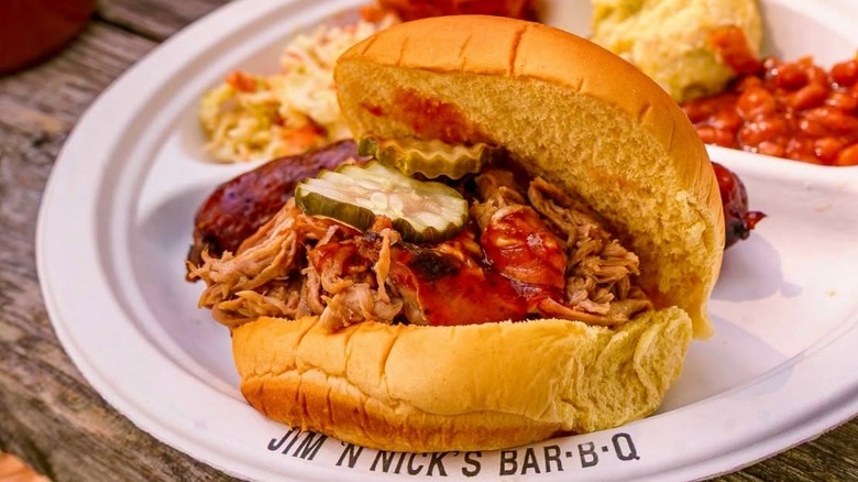 A barbecue pulled pork sandwich with hot sauce and pickles sits on a plate with classic Southern sides, including baked beans and biscuits.