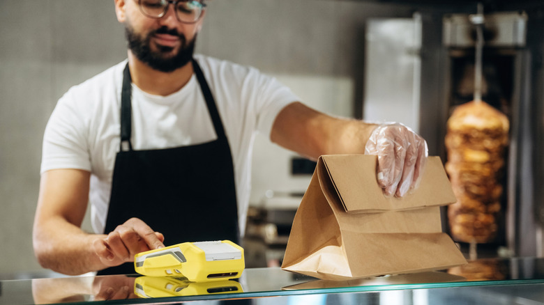 A restaurant employee hands off a bag of takeout while ringing up the total for the customer.