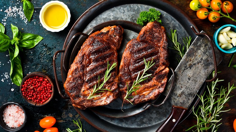 Two grilled ribeye steaks on a steel pan, topped with rosemary sprigs with a butcher's knife next to them, surrounded by accoutrements and seasonings: olive oil, salt, basil, peppercorns, rosemary, peeled garlic, and cherry tomatoes.