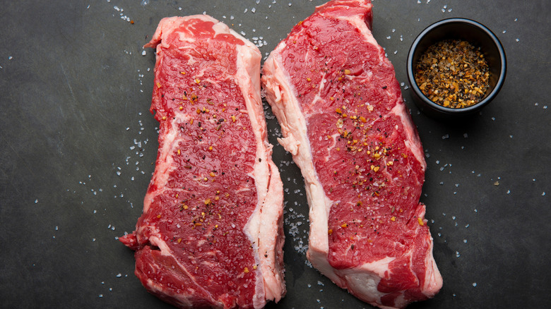 Raw strip loin steaks on black surface with salt and spices