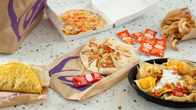 Taco Bell combination of Crunchwrap Supreme, hard shell taco, nachos, cinnamon twists, and Mexican pizza, with Taco Bell bag showing, all on a light table.
