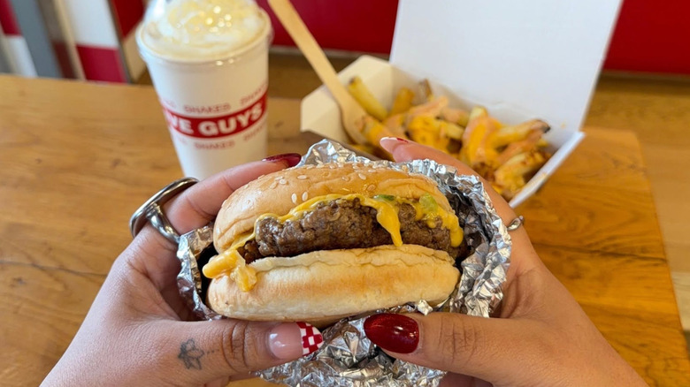 Someone holds up a Five Guys burger in front of a milkshake and cheesy fries.