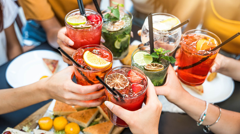 Seven drinks being held and clinked together over a table with plates and food