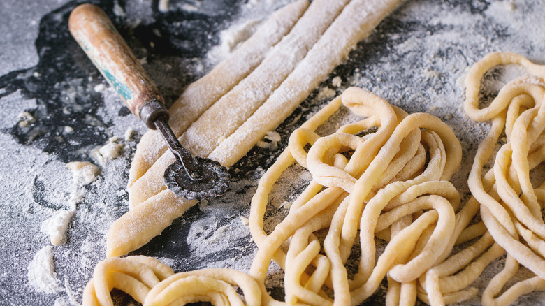 Fresh spaghetti-like pici pasta, some rolled and some still flat,  on a black floured surface with a pastry wheel cutter