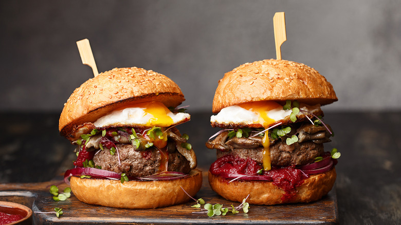 Two homemade beef burgers with mushrooms, micro greens, red onion, fried eggs, and beet sauce on wooden cutting board.