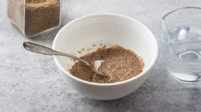 A white bowl containing a flax egg substitute mixture sits on a gray counter, with a measuring cup of water and a glass jar of flax meal sitting adjacent