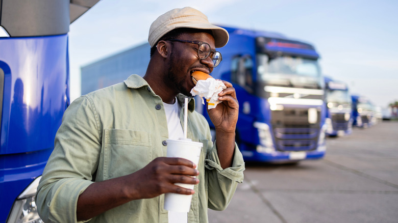A truck driver eats a burger and holds a soda near his big rig