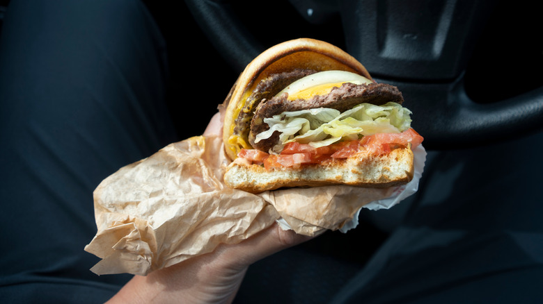 A hand is shown holding a fast-food burger wrapped in parchment paper