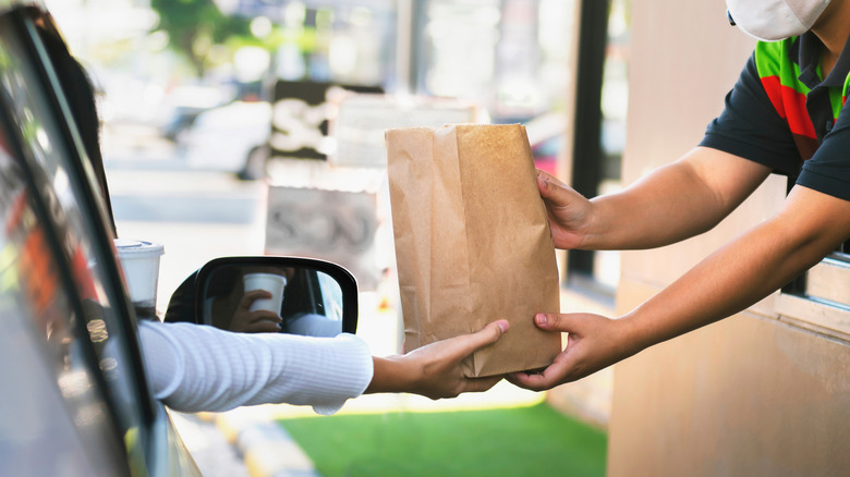 Person in car receiving brown paper bag from drive-through window at fast food spot.