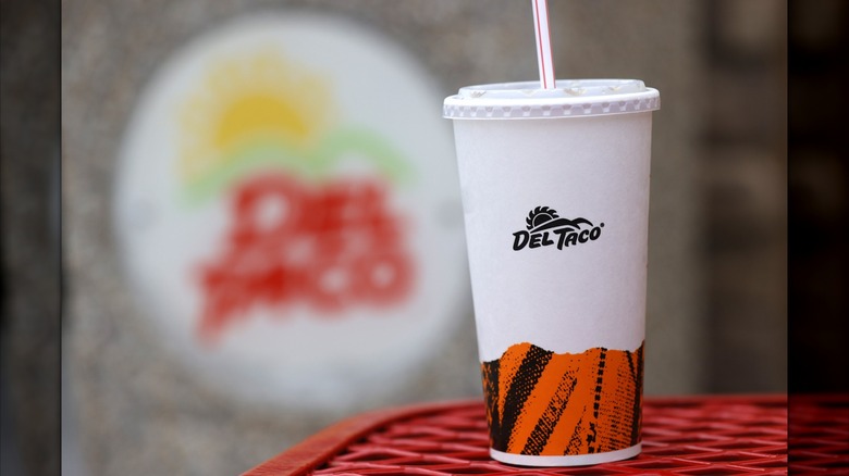 A Del Taco soda cup sits on a red table, with the Del Taco sign blurred out in the background