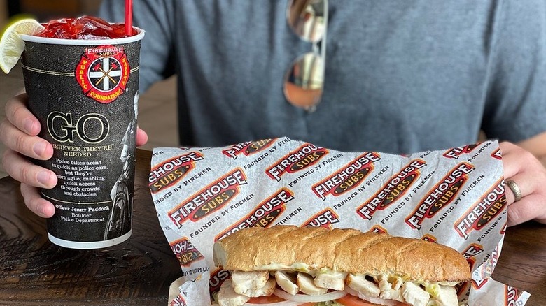 A diner holds a Firehouse Subs drink cup next to an unwrapped sandwich