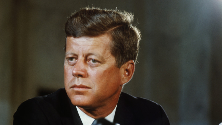 U.S. President John F. Kennedy looks to the side while seated in front of a mic