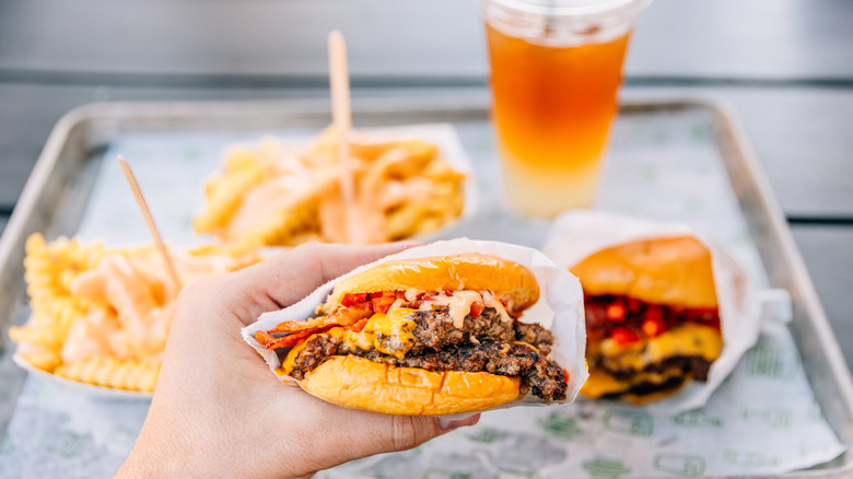 Person holding a burger in front of a tray of fast food