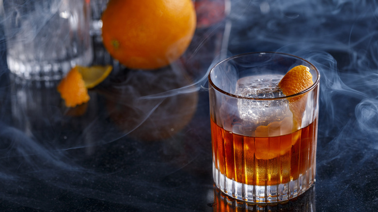 Bourbon in a rocks glass with orange peel. Smoke surrounding the glass, with full orange and additional rocks glasses blurred in the background. All on a dark table.