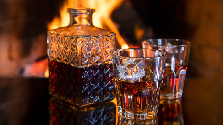 Two rocks glasses of bourbon near a decanter half-way filled with bourbon. All on a table with a fire burning in the background.