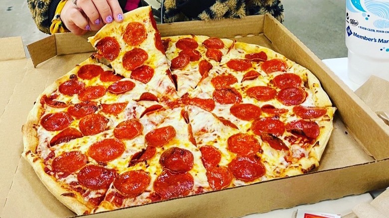 A woman holds up a piece of Sam's Club pizza from the rest of the pie in its box