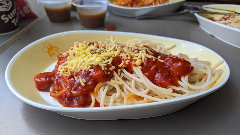 Spaghetti with a red sauce with hot dog slices and shredded yellow cheese in a yellow and white plate