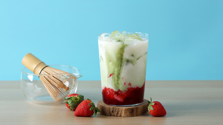 An iced strawberry matcha latte with fresh strawberries on the table next to a matcha whisk
