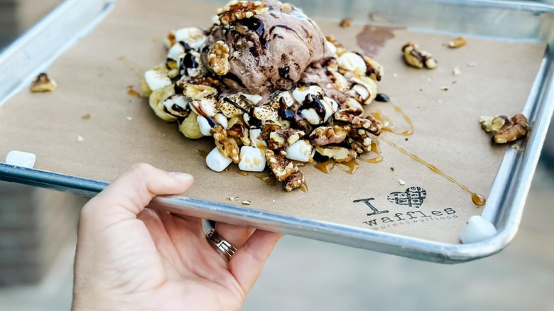 Someone holds a rocky-road themed waffle, topped with chocolate ice cream, mini marshmallows, nuts, and chocolate sauce