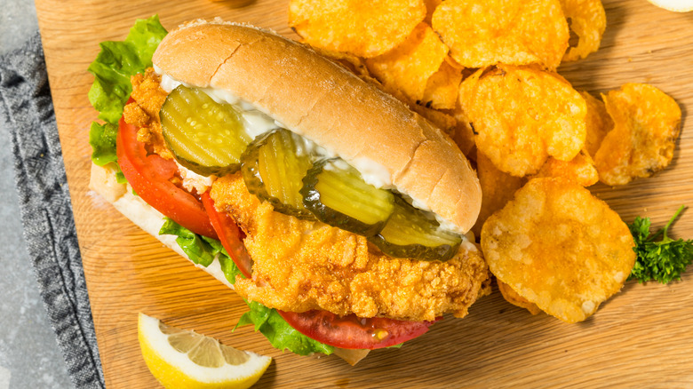 A fried fish sandwich sits on a cutting board next to a pile of chips.