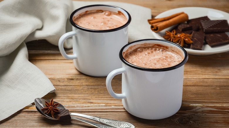 Two cups of hot cocoa on a wooden table with bars of dark chocolate, cloves, and cinnamon.