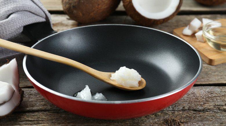 Coconut oil in a red pan with a wooden spoon surrounded by fresh coconut