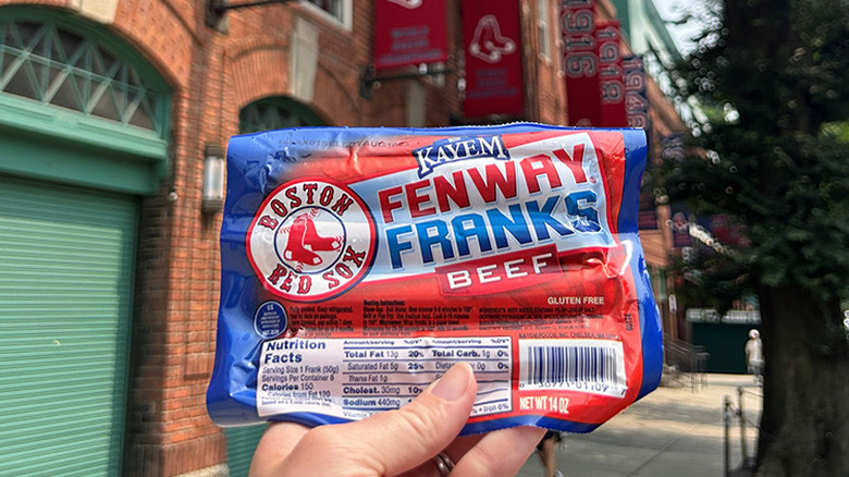 A hand holds up a package of Kayem Fenway Franks in an outdoor setting in Boston